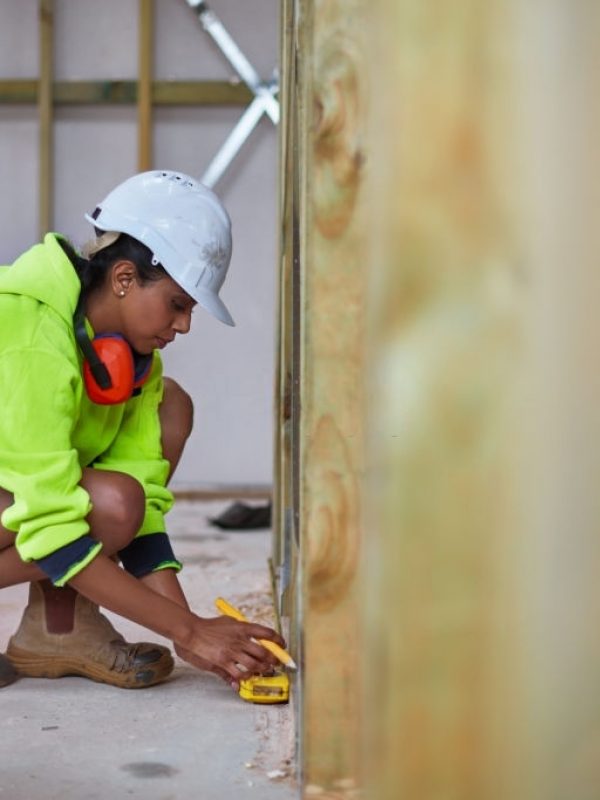 Full length of construction worker using tape measure while working. Female engineer is in reflective clothing. She is crouching on floor at construction site.